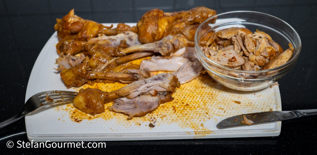 A cutting board with chicken drumsticks and shredded chicken pieces, showcasing the preparation process for Khao Soi.