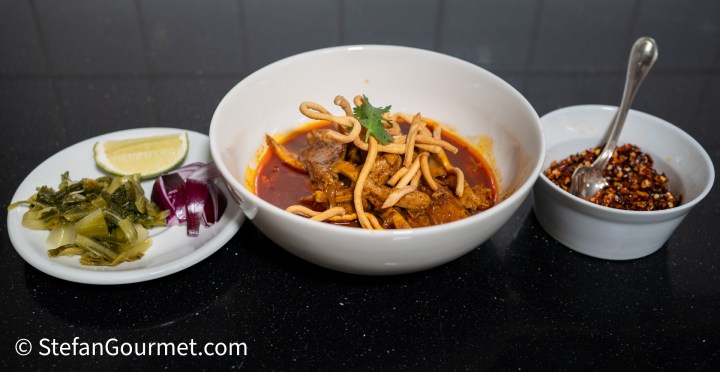 A bowl of Khao Soi featuring rich curry soup with chicken, topped with crispy fried noodles, accompanied by a plate of lime wedges, sliced red onion, and pickled mustard greens.