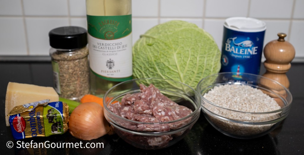 Ingredients for risotto including cabbage, sausage, rice, carrot, onion, garlic, white wine, Parmigiano cheese, and seasonings arranged on a countertop.