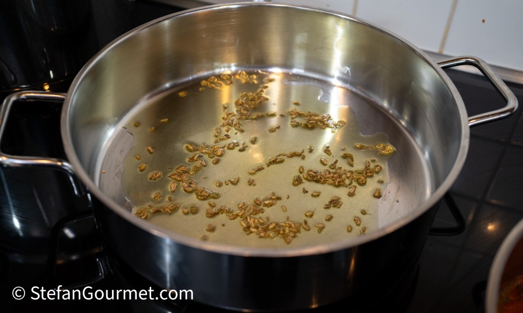 A stainless steel pan on a stove, containing oil and toasted fennel seeds.