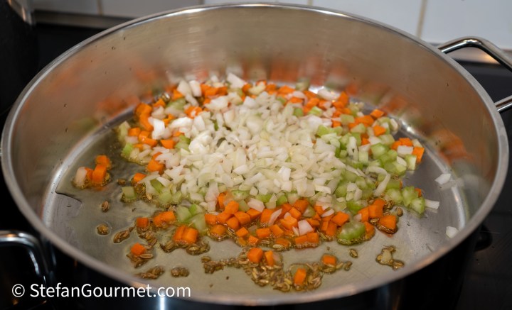 A large skillet with sautéed finely diced onion, carrot, and celery, with fennel seeds visible in the bottom.