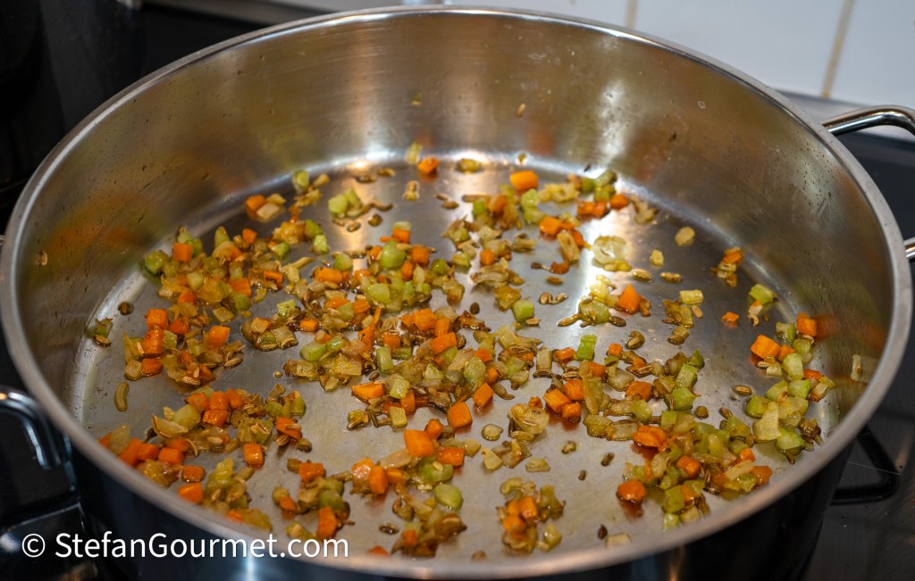 Finely diced carrots, celery, and onions sautéing in a large stainless steel pan, with some fennel seeds visible on the bottom.