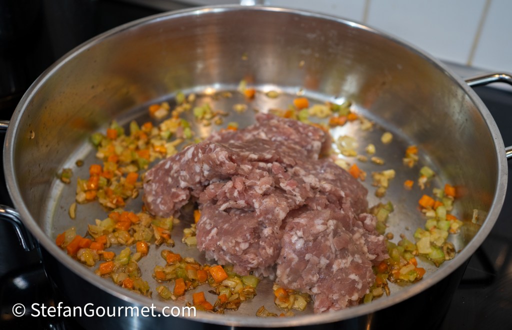 Ground Italian sausage placed in a large pan with sautéed diced vegetables including carrot, onion, and celery.