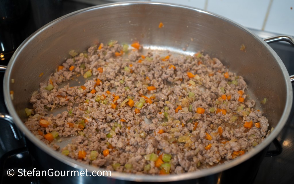 A sauté pan with cooked Italian sausage crumbles mixed with diced vegetables including carrots and celery, situated on a stovetop.