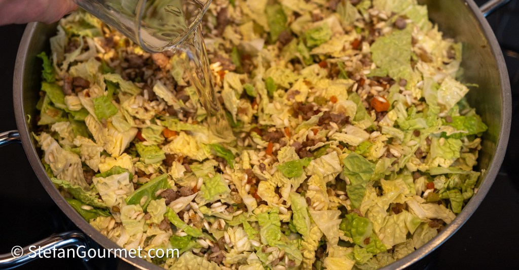 A hand pouring liquid into a large pan filled with shredded savoy cabbage, Italian sausage, and rice, with a mix of ingredients visible.