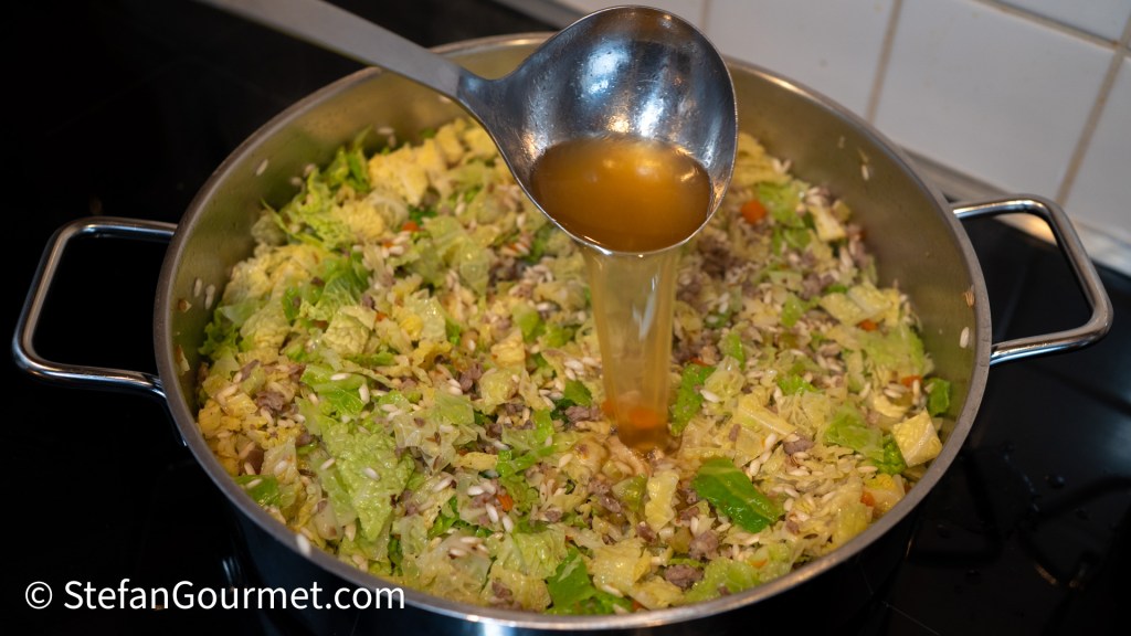 A large pot filled with a mix of shredded savoy cabbage, risotto rice, and meat, with a ladle pouring hot stock into the mixture.