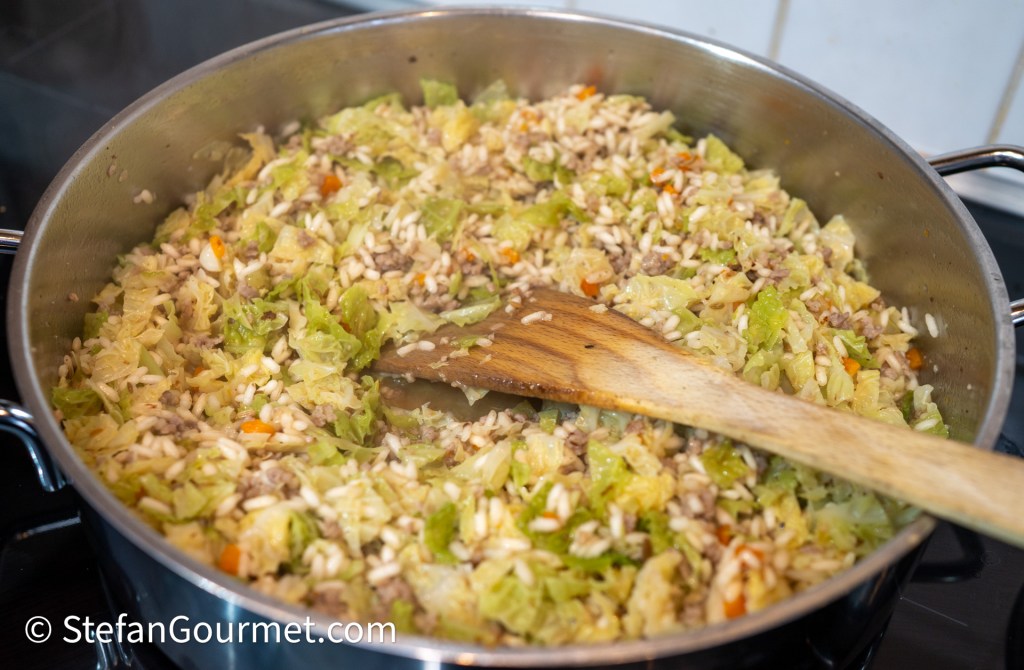 A pot filled with risotto rice, shredded savoy cabbage, and minced vegetables cooking together, with a wooden spoon resting in the mixture.