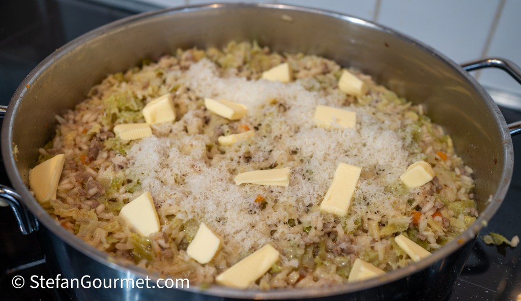 A pot of creamy risotto with shredded savoy cabbage, chunks of butter, and grated cheese on top, cooking on the stovetop.