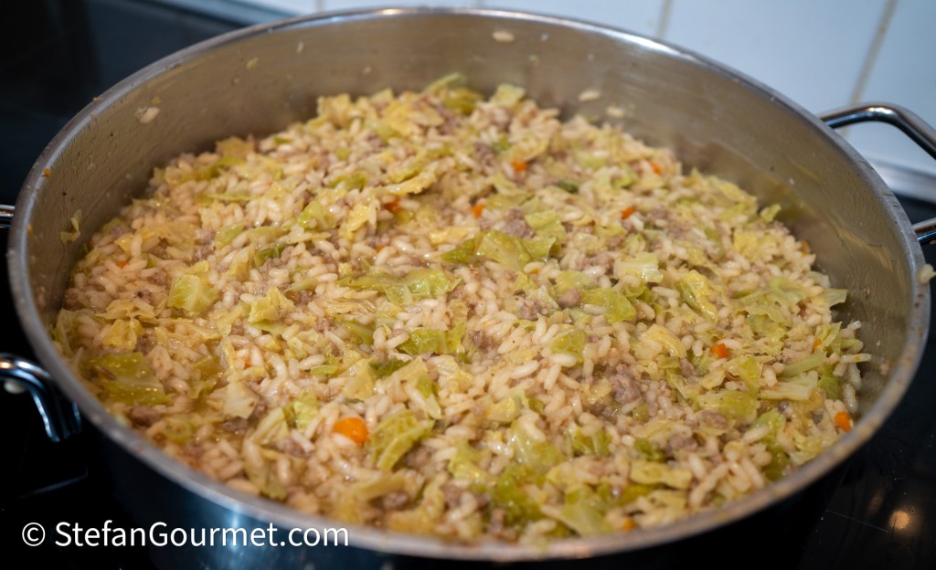 A pot filled with savory risotto containing rice, shredded savoy cabbage, and pieces of Italian sausage, simmering on a stovetop.