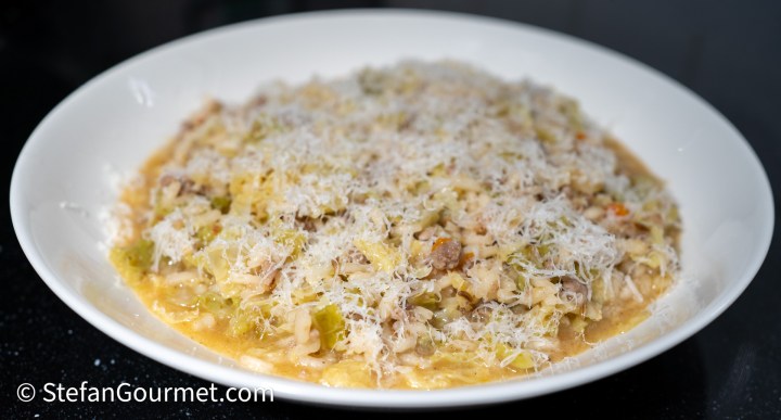 A plate of creamy risotto topped with grated cheese, featuring bits of Italian sausage and shredded savoy cabbage, served on a dark surface.