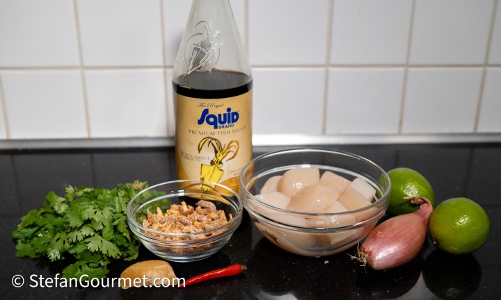 Ingredients for scallop ceviche featuring raw scallops, fish sauce, cilantro, dried shrimp, lime, shallots, and chili laid out on a black countertop.