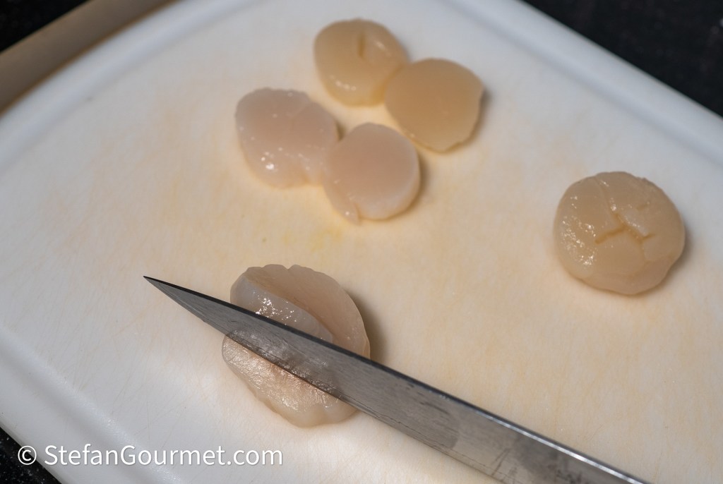 A close-up of a chef's knife slicing through raw scallops on a white cutting board.