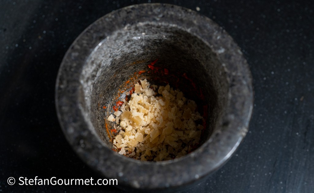 A close-up view of a traditional mortar and pestle containing a mixture of ground dried shrimp and red chili, showcasing the textured ingredients inside.