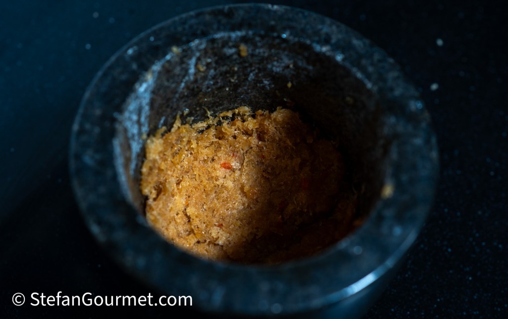 A close-up of a mortar filled with a coarse paste made from dried shrimp, chili, and palm sugar, set against a dark background.