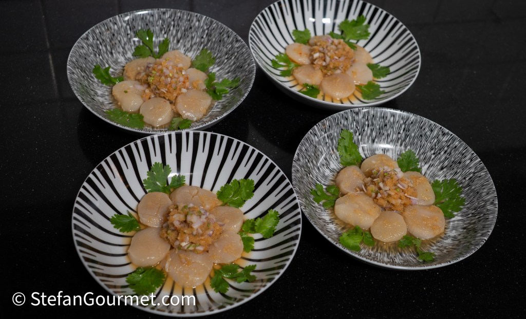 Four plates of scallop ceviche arranged in a flower pattern, garnished with minced shrimp and cilantro leaves, set on a black countertop.