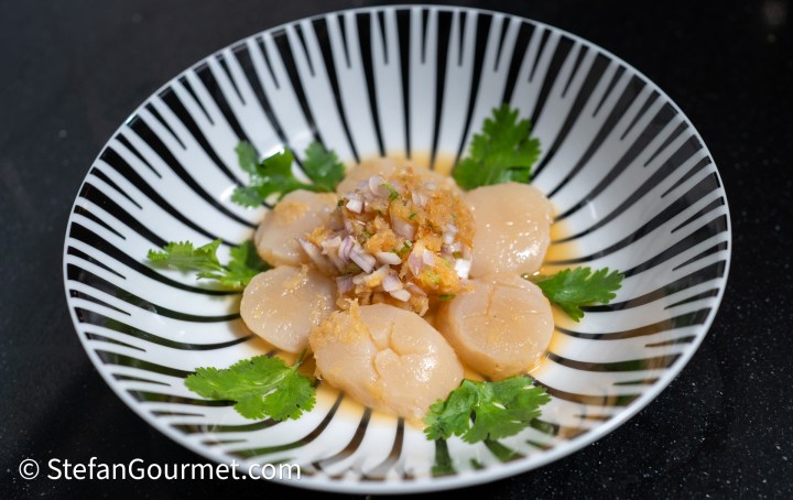 A decorative bowl filled with scallop ceviche arranged in a flower pattern, topped with a mixture of shallots and dressing, garnished with fresh cilantro leaves.