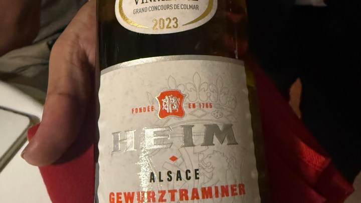 A close-up of a bottle of Alsace Gewürztraminer wine, held by a person, featuring its label with the name Heim and vintage year 2023.