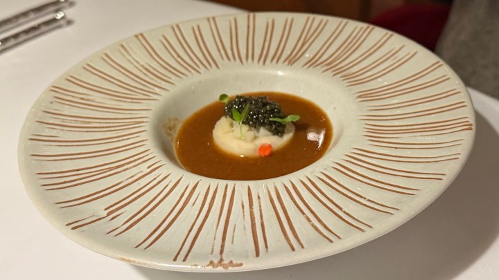 A sophisticated dish presented in a decorative plate, featuring a creamy base topped with dark caviar and garnished with green herbs, surrounded by a rich brown sauce.