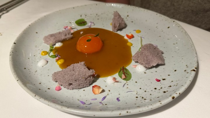 A plate featuring a modern interpretation of a Thai dessert, showcasing a vibrant orange sauce, a bright egg yolk, and delicate purple cake pieces, garnished with edible flowers and colorful sauces.