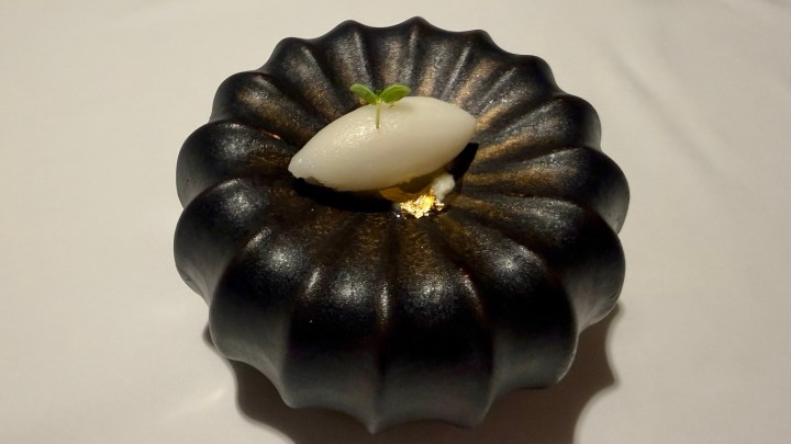 A black, fluted dessert dish with a white sorbet topped with a small green mint leaf, placed on a white tablecloth.