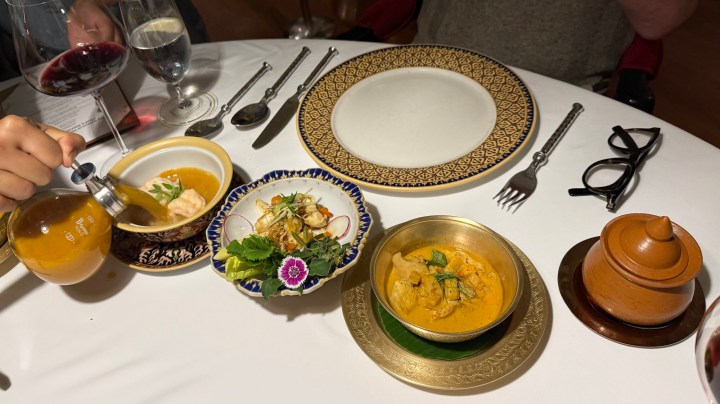 A fine dining table set with a decorative plate, various Thai dishes including a curry, a salad, and a soup, and a hand pouring broth from a glass container.