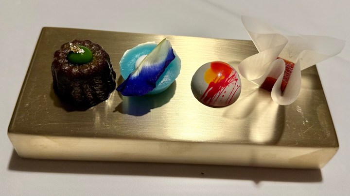 A beautifully arranged selection of colorful petit fours set on a gold board, featuring intricate desserts like a chocolate canelé, a blue jelly with a flower petal, a decorative round confection, and another sweet wrapped in a delicate paper.