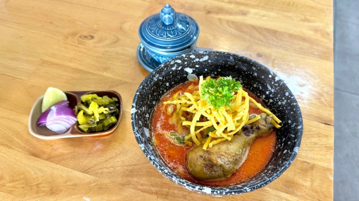 A bowl of Khao Soi featuring rich curry noodle soup with a chicken drumstick, topped with crispy fried noodles and garnished with cilantro. Accompanied by a side of pickled mustard greens, sliced red onion, and a lime wedge.