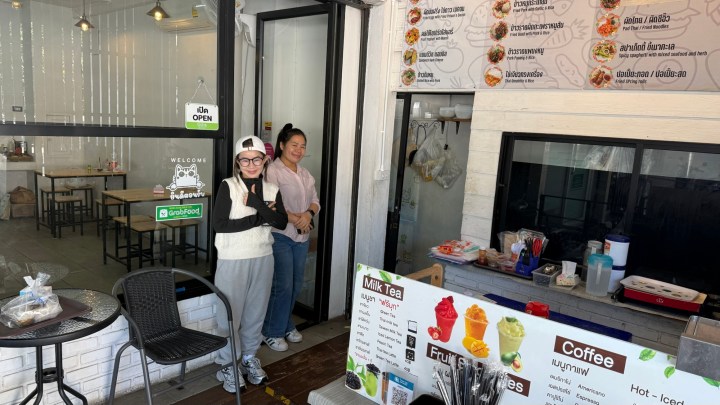 Two women smiling at the entrance of a small Thai restaurant, with a menu displayed on a counter nearby.