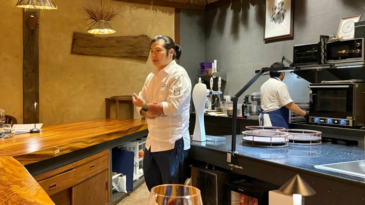 A chef in a white jacket standing at a wooden counter in a restaurant kitchen, explaining a dish to guests.
