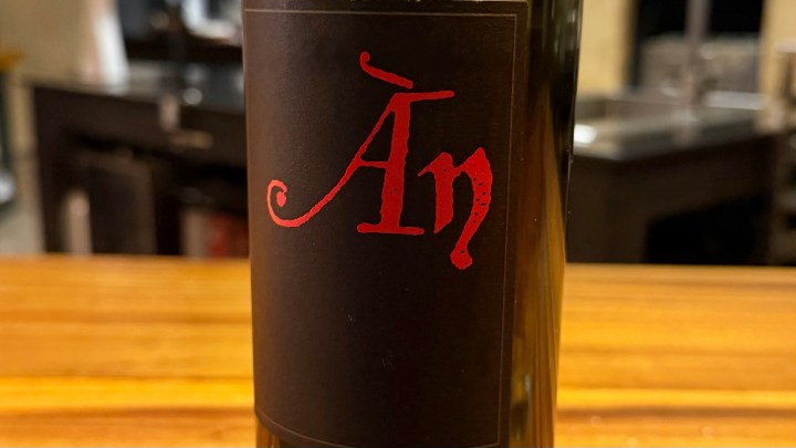 A close-up of a wine bottle featuring a black label with red text that reads 'Án', placed on a wooden surface.