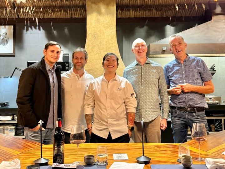 A group of five individuals, including a chef, standing together in a well-decorated restaurant with a stylish wooden table and modern kitchen backdrop.