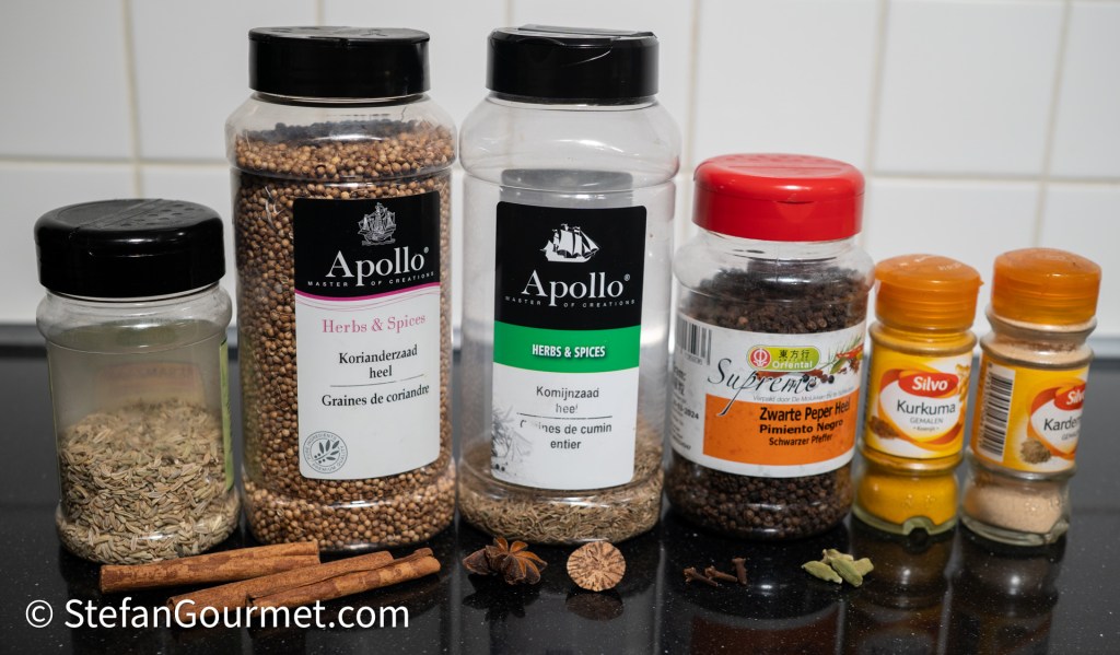 A variety of spices used for cooking, including coriander seeds, cumin seeds, black pepper, turmeric, cinnamon sticks, star anise, and ground cardamom, displayed in glass jars on a black countertop.