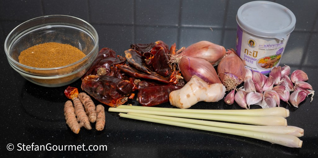 Ingredients for Gaeng Hung Lay curry, including curry powder, dried red chilies, shallots, garlic, lemongrass, turmeric root, and shrimp paste arranged on a dark countertop.