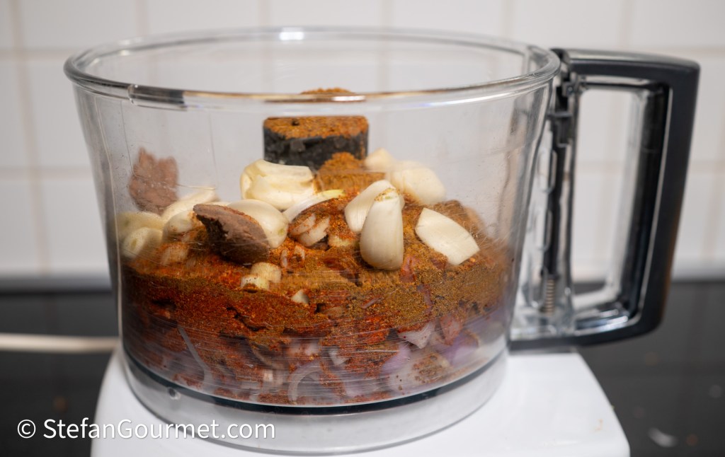 A food processor filled with ingredients for curry paste, including spices, garlic, and shallots, ready for blending.