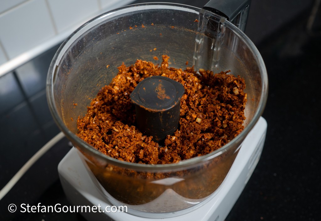 A food processor containing a blend of spices and ingredients for making curry paste, with a dark reddish-brown color and a spatula placed in the center.