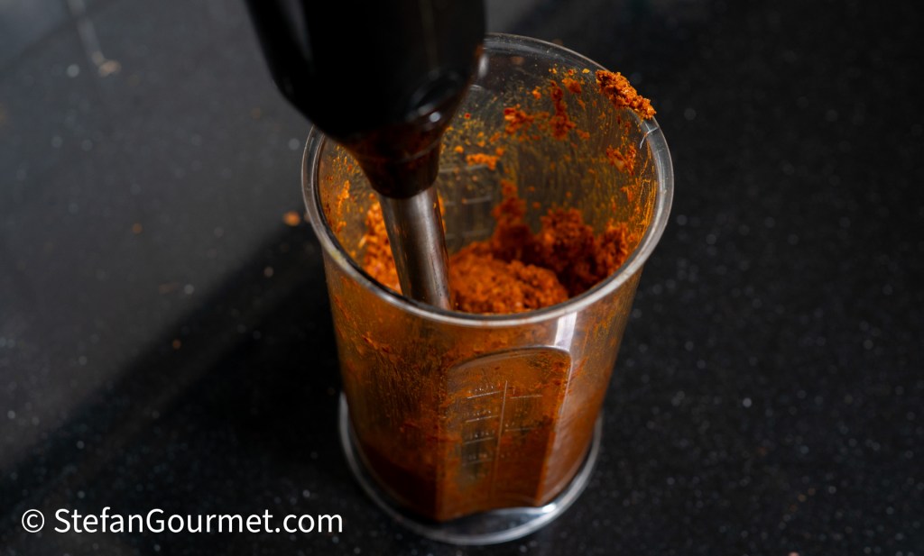 An immersion blender mixing red curry paste in a measuring cup on a black countertop.
