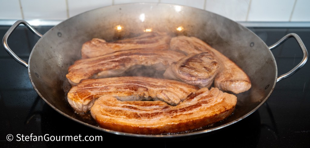 Slices of browned pork belly cooking in a shallow metal pan with steam rising.