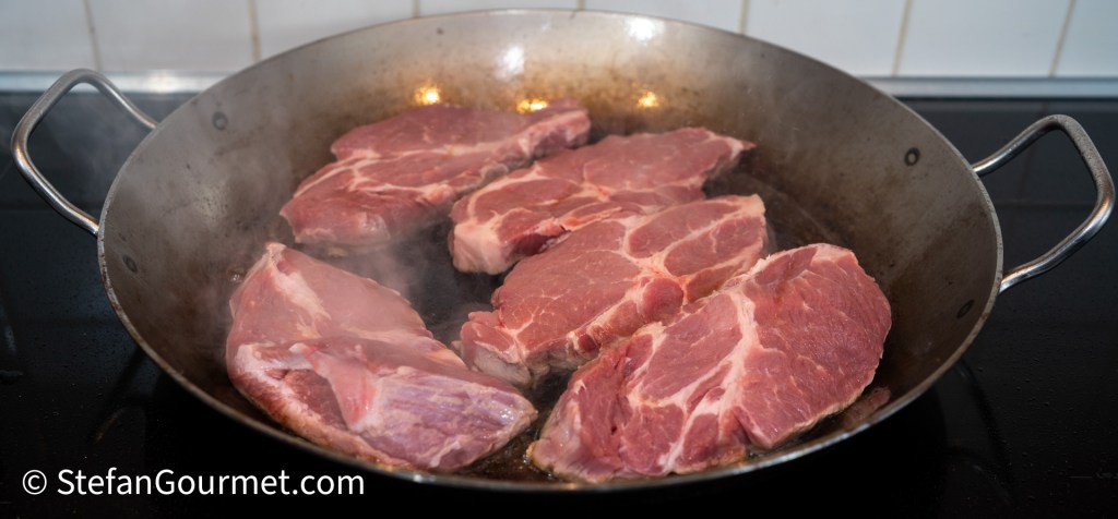 Pork slices browning in a large pan on a stovetop.