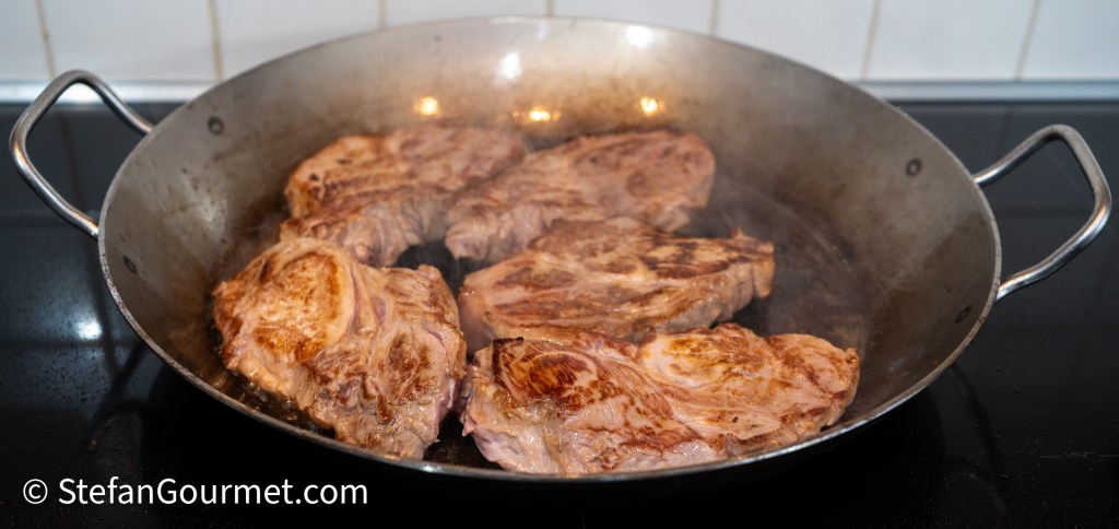 Seared pork belly slices cooking in a large metal pan on a stovetop.