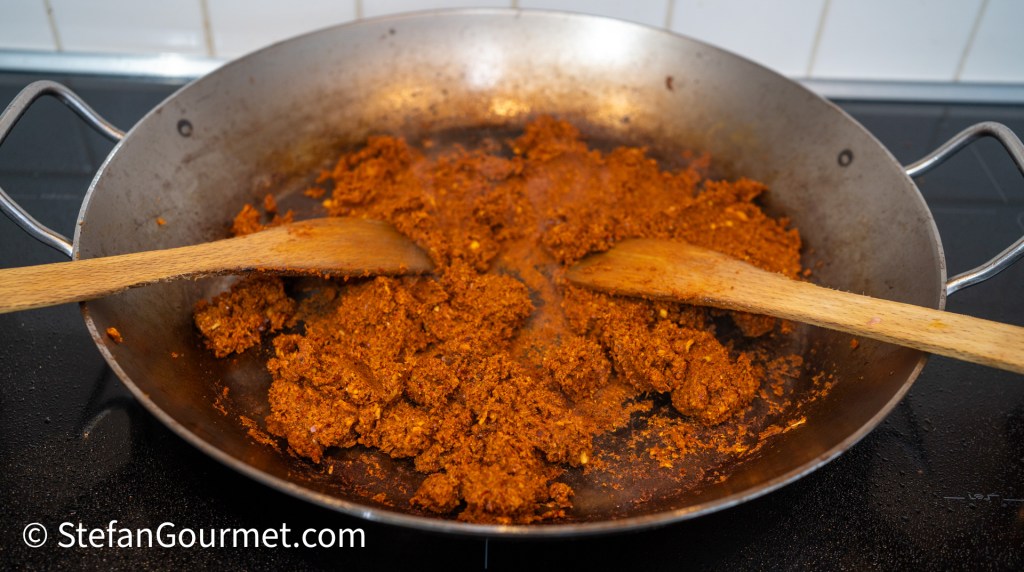 A wok with a reddish-brown curry paste being stirred with two wooden spoons, on a stovetop.