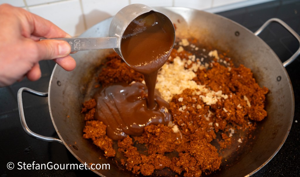 A hand pouring sauce into a pan containing ginger and red curry paste.