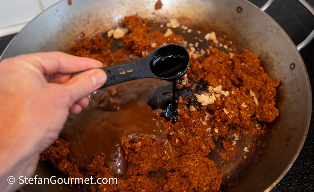 A close-up of a hand holding a measuring spoon pouring dark liquid into a pan containing a mixture of spices and ingredients for curry preparation.