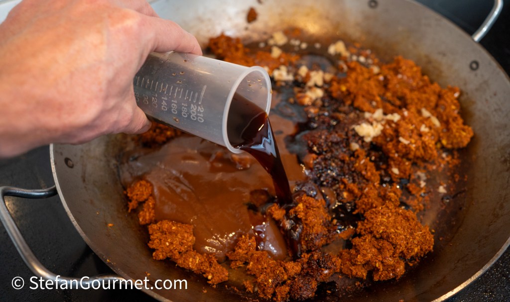 A person pouring a dark liquid from a measuring cup into a pan containing spices and a thick paste, with a focus on the mixing process.