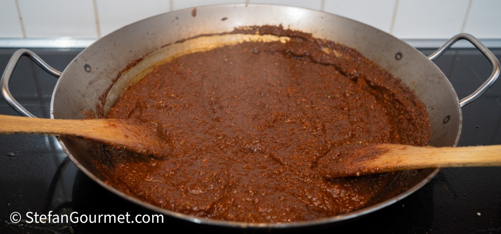 A large pan with curry paste simmering, featuring a rich brown color and a wooden spoon stirring the mixture.