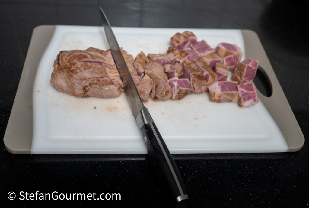 Sliced pork belly on a cutting board with a knife, showing both whole and cubed pieces.