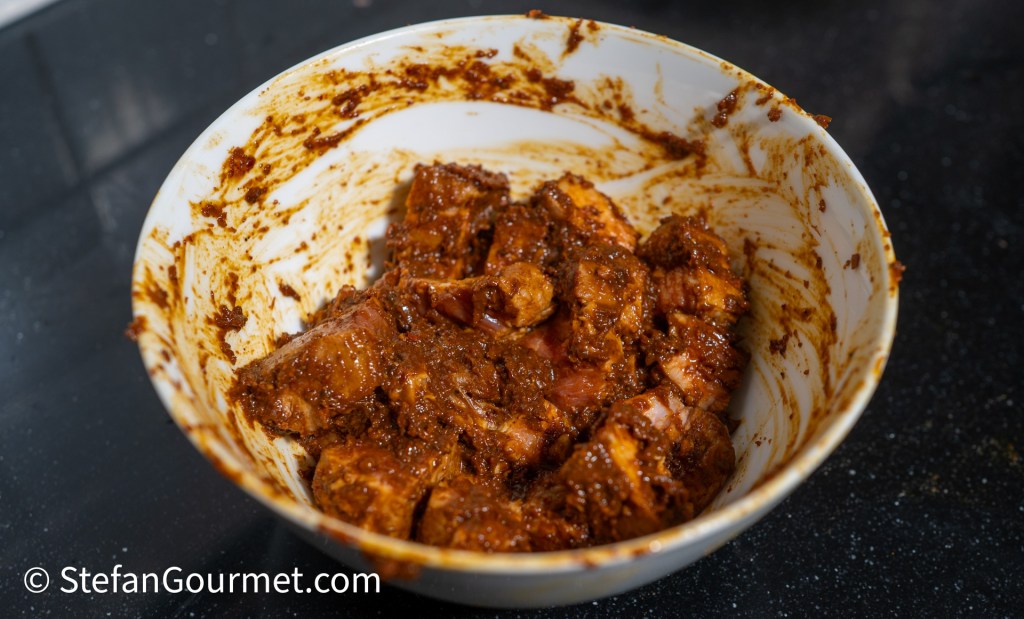 A bowl containing marinated pork pieces coated in a rich, dark curry paste, set against a dark countertop.