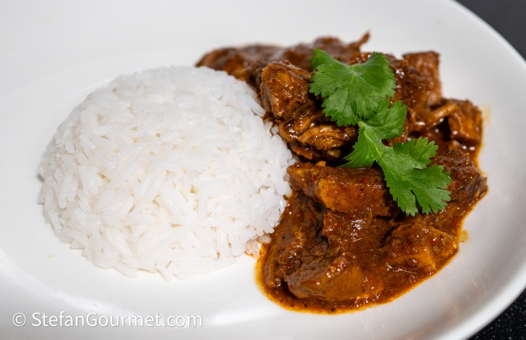 A serving of Gaeng Hung Lay curry with pieces of pork and a side of steamed jasmine rice garnished with fresh cilantro.