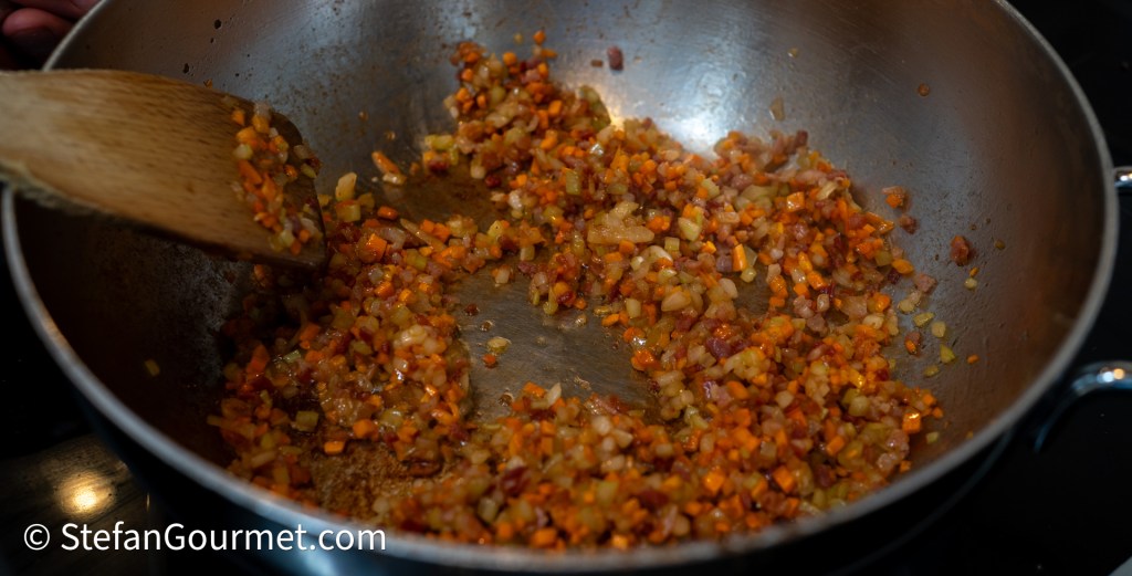 A sauté pan with finely chopped vegetables and pancetta being cooked, showcasing a blend of diced carrots, onions, and celery.