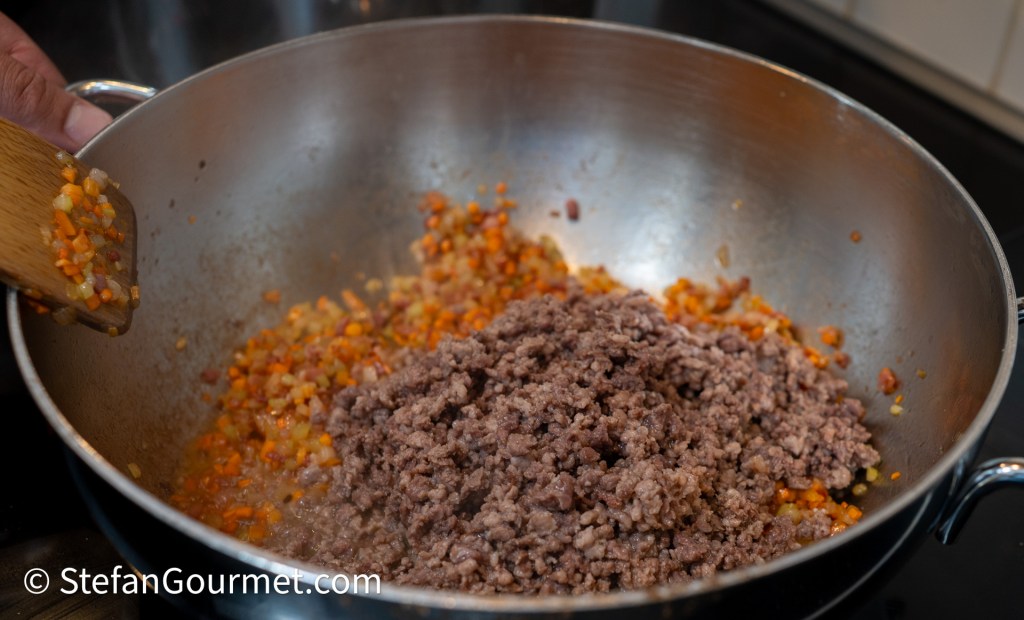 A pot with sautéed vegetables and ground meat cooking together, showing a wooden spatula stirring the mixture.