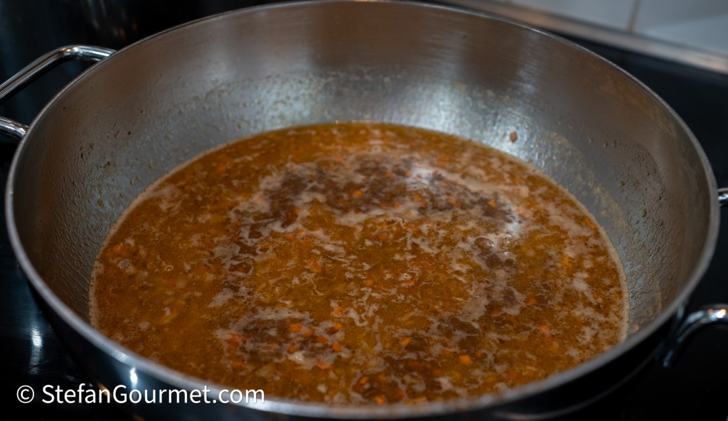 A close-up view of a pot containing a simmering meat sauce with visible herbs and bits of vegetables.